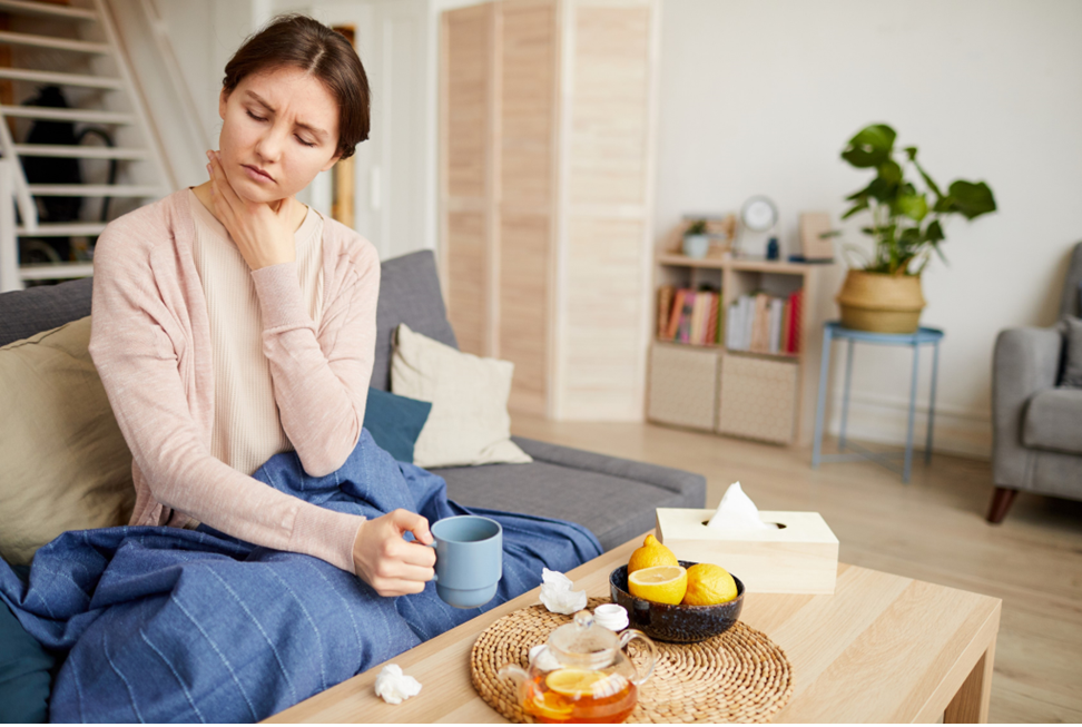 Woman holding her throat in discomfort by sitting in sofa with cup in other hand.