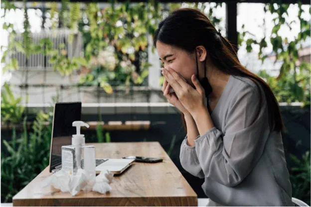 A_woman_with_a_cough_and_cold_at_her_desk_blowing_nose.webp